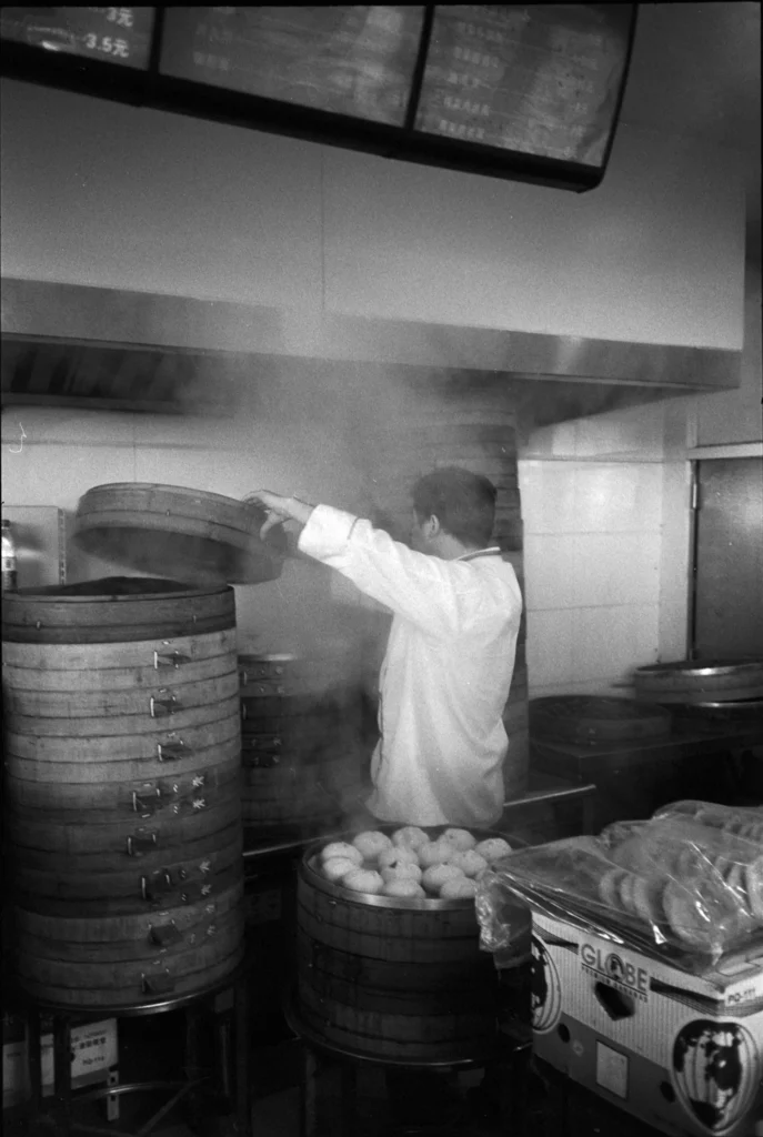 Cuisinier de dos soulevant le couvercle d'un panier vapeur en bambou dans une echoppe de baozi, vapeur et paniers empiles