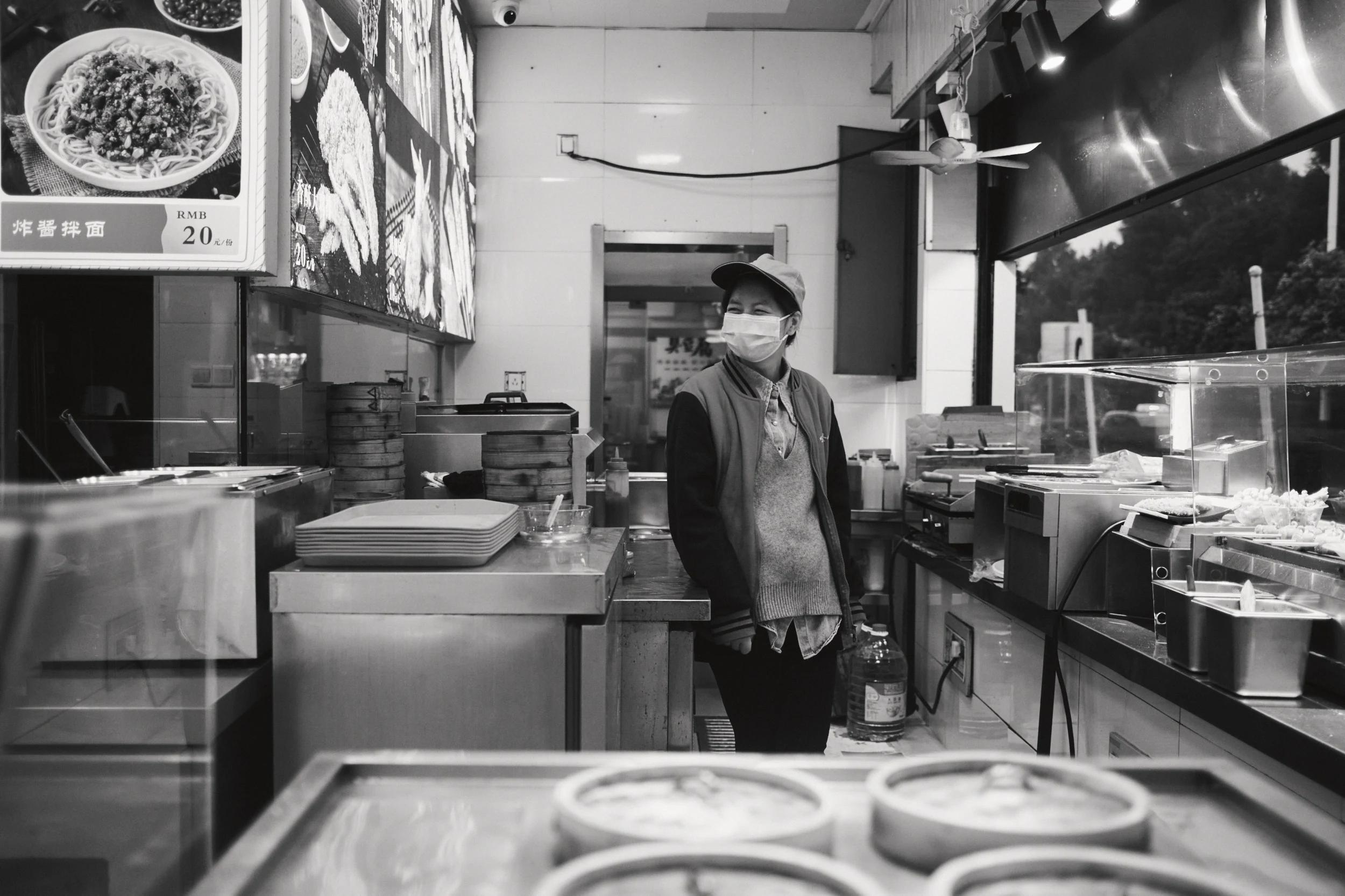 Employee coiffee d'une casquette et masquee derriere le comptoir d'une echoppe de nouilles et raviolis vapeur, menus photos au mur