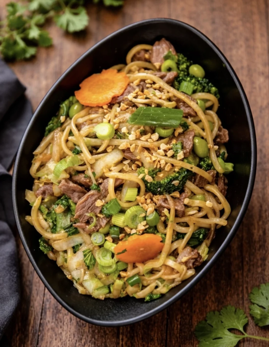 Bol noir de nouilles sautees au boeuf avec brocoli, edamame, carotte et cacahuetes sur une table en bois