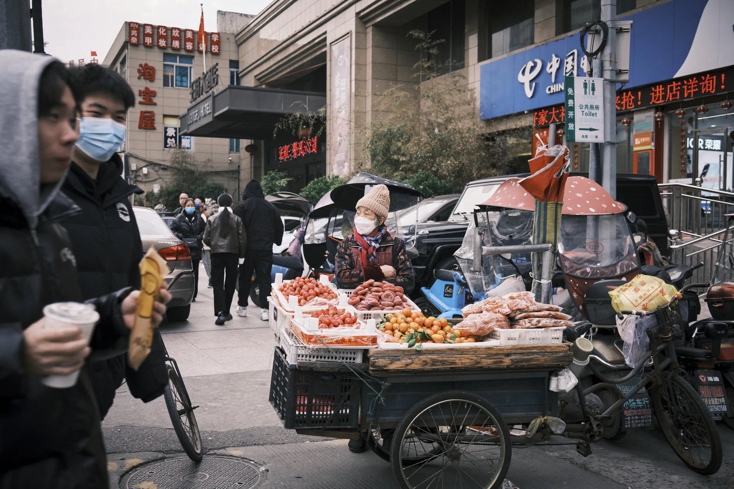 Vendeuse ambulante agee vendant fraises, oranges et saucisses depuis un triporteur dans une rue commercante, passants au premier plan